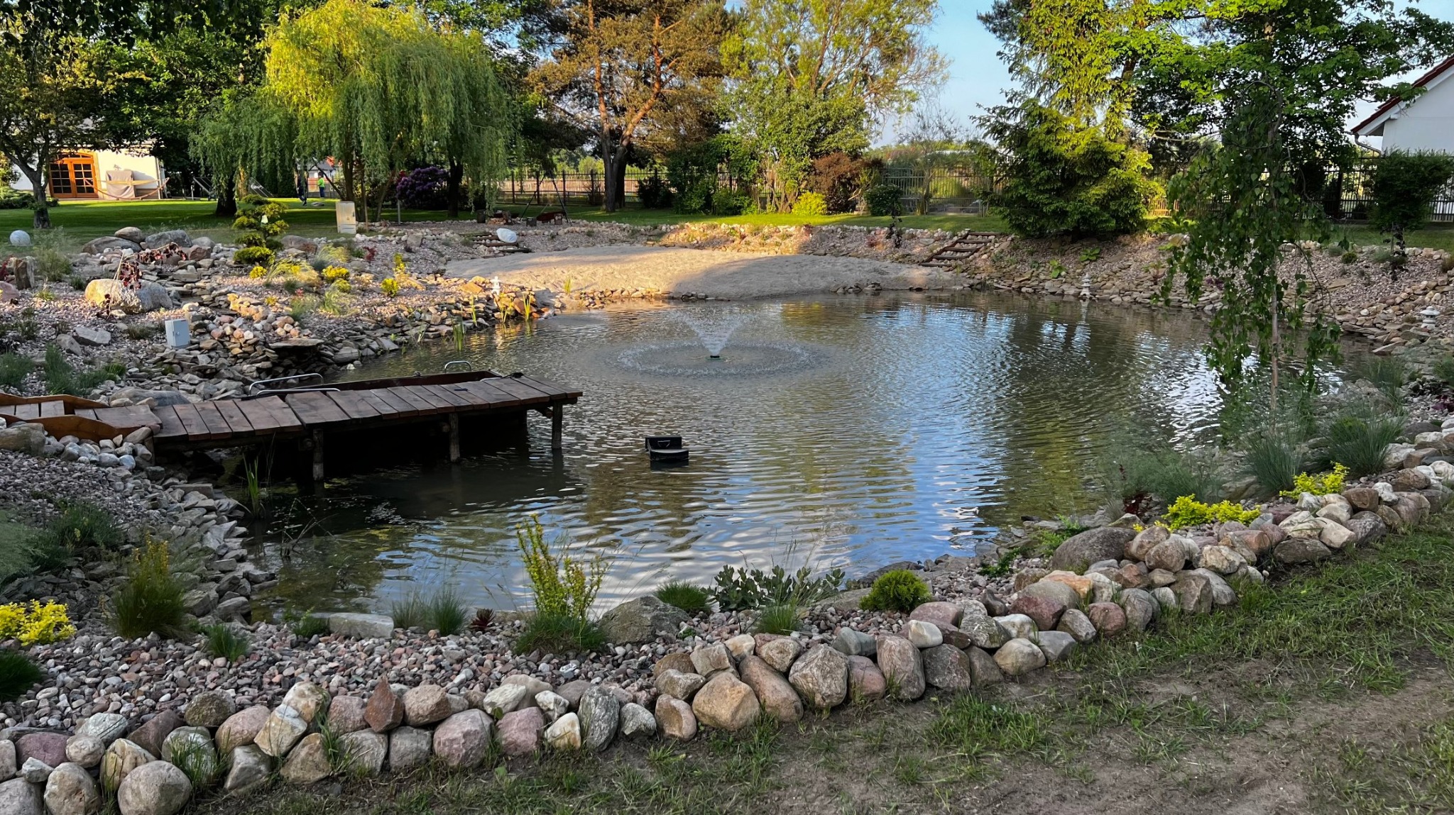 Oczko wodne z fontanną i drewnianym pomostem, otoczone kamieniami i zielenią. Uroku dodają drzewa i rośliny w tle, tworząc sielski krajobraz.