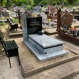 Granitowy nagrobek w Pyrzycach z czarną tablicą i szarym obramowaniem, otoczony innymi grobami i zielenią cmentarza. Obok ławka.
