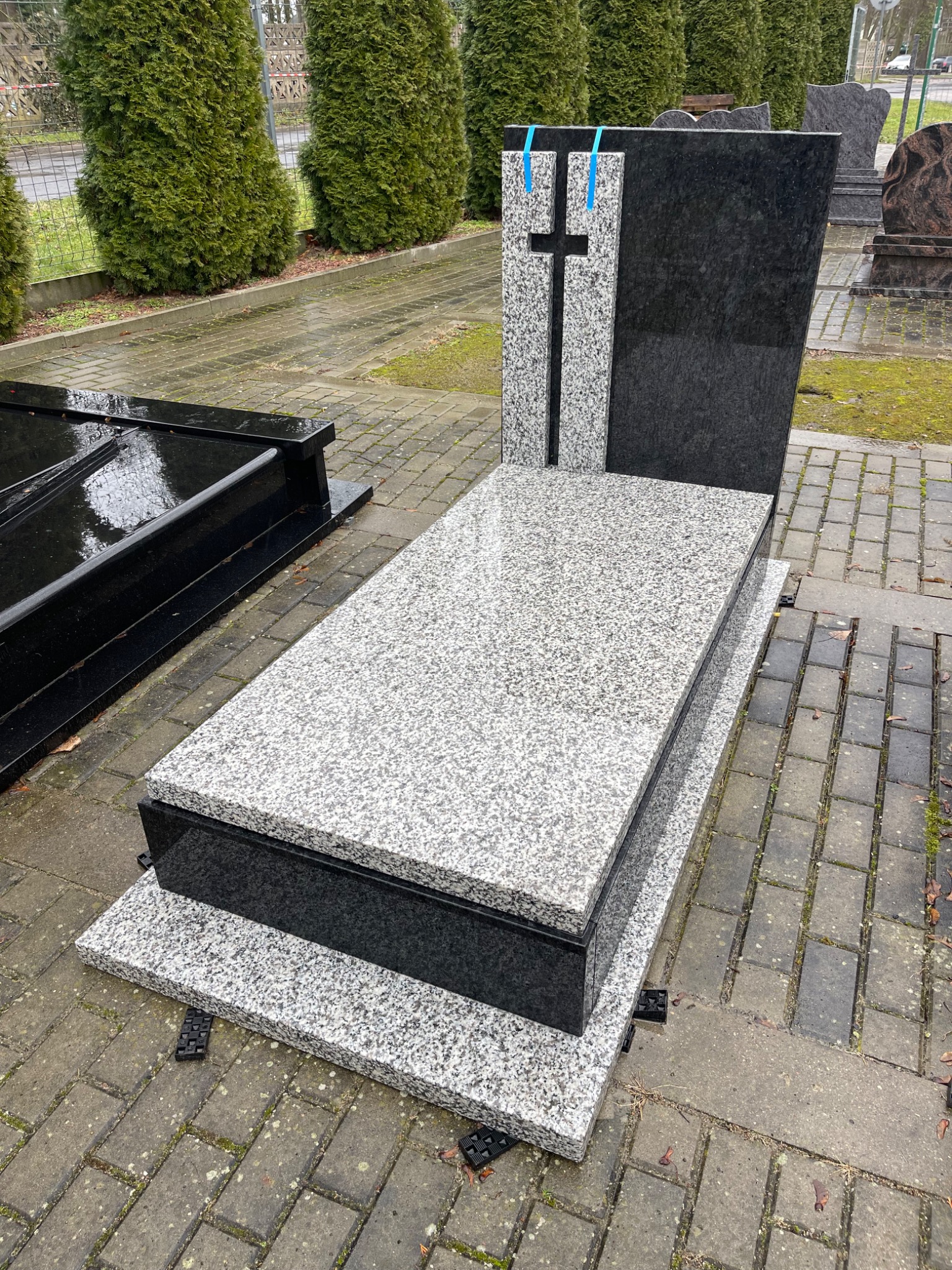 Granitowy nagrobek z krzyżem, jasnoszary blat i ciemne elementy, ustawiony na kostce brukowej. Widok z góry, w tle zieleń.