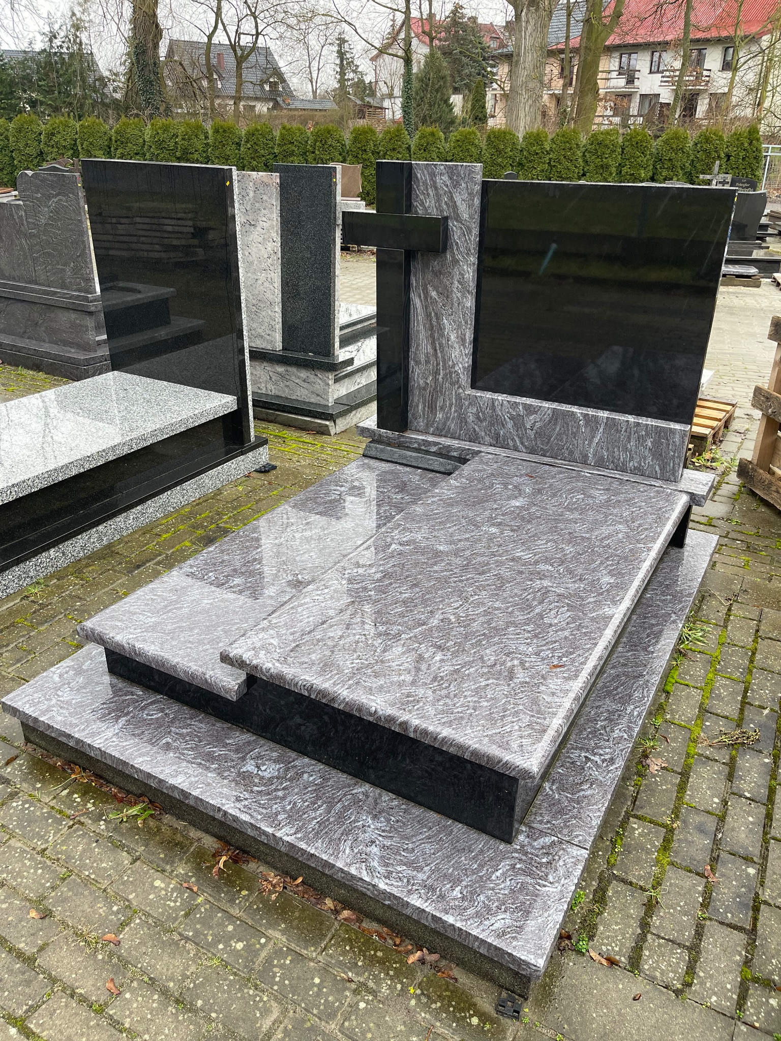 Granitowy nagrobek z krzyżem, w odcieniach szarości i czerni, ustawiony na cmentarzu. Widoczne inne nagrobki w tle, tło stanowią drzewa i budynki.