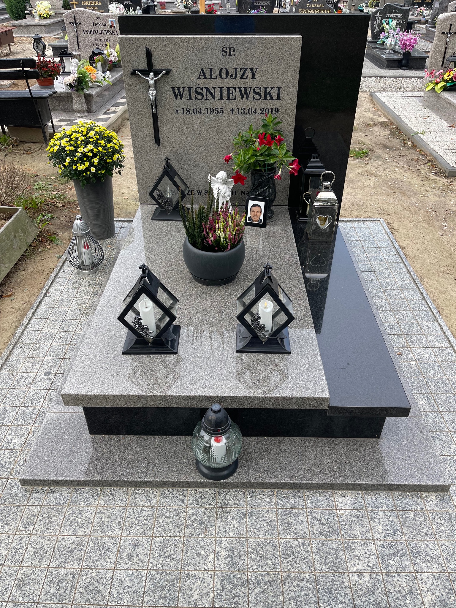 Granitowy nagrobek z czarnym akcentem, ozdobiony kwiatami, lampionami i zdjęciem. Całość umieszczona na kostce brukowej. Widok z góry.