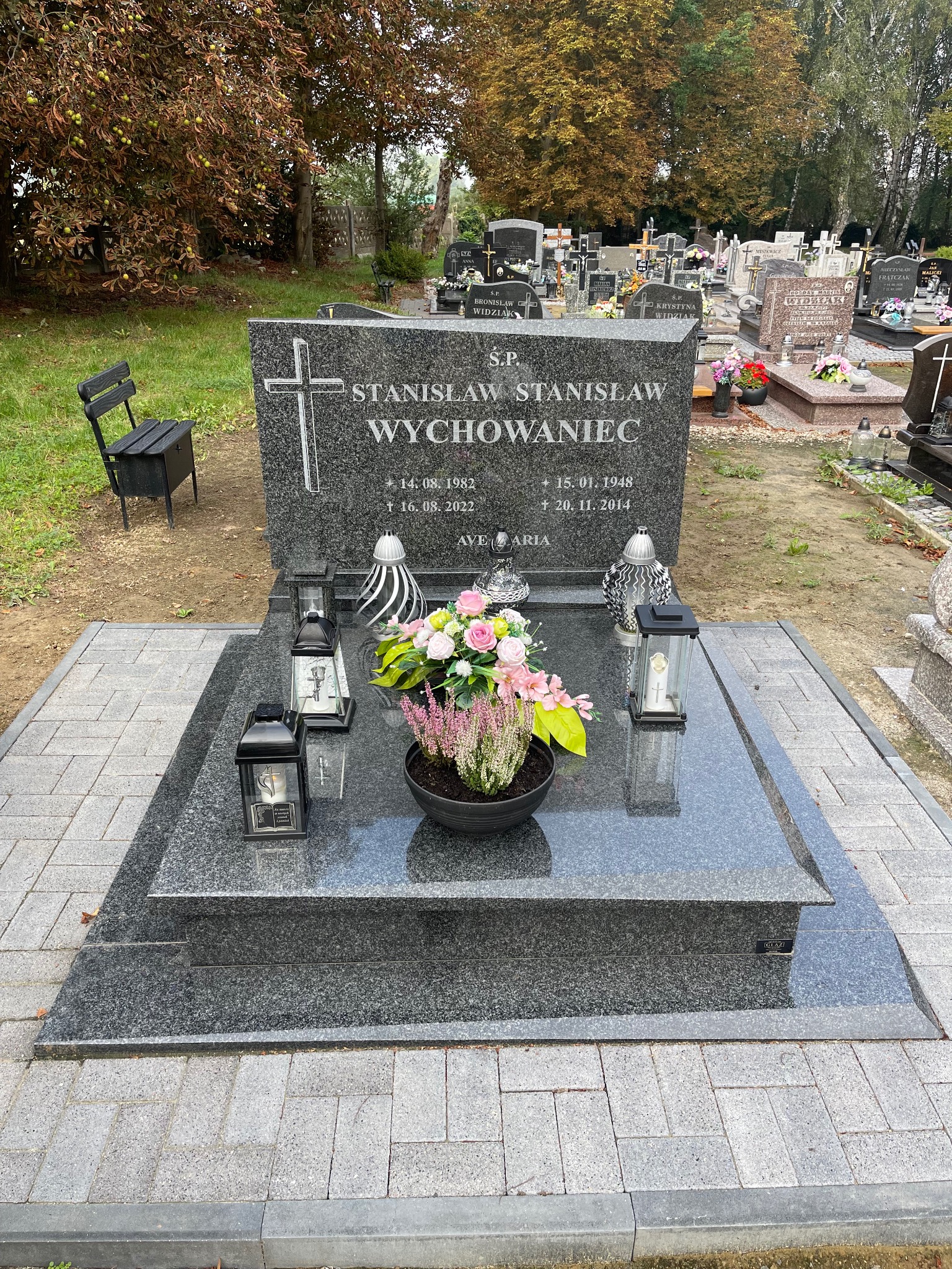 Granitowy nagrobek z krzyżem i inskrypcją, otoczony lampionami i kwiatami w donicy. Ułożona kostka granitowa. Widok na cmentarz w jesiennej scenerii.