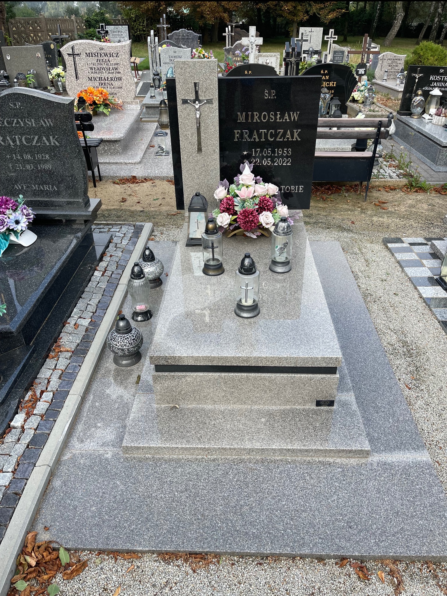 Granitowy nagrobek z kwiatami i lampionami na cmentarzu. Jasnoszara płyta nagrobna z ciemniejszym napisem. Otoczenie żwirowe.