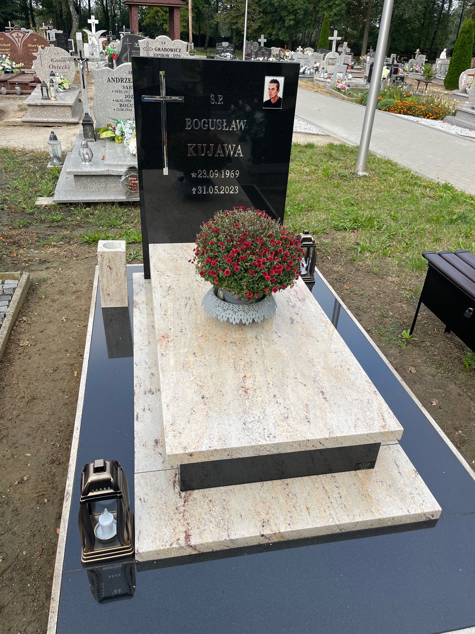Czarny, polerowany nagrobek z jasnym blatem i kwiatami na cmentarzu. Portret osoby w rogu. Napis: B. Kujawa, daty urodzin i śmierci. Lampion.