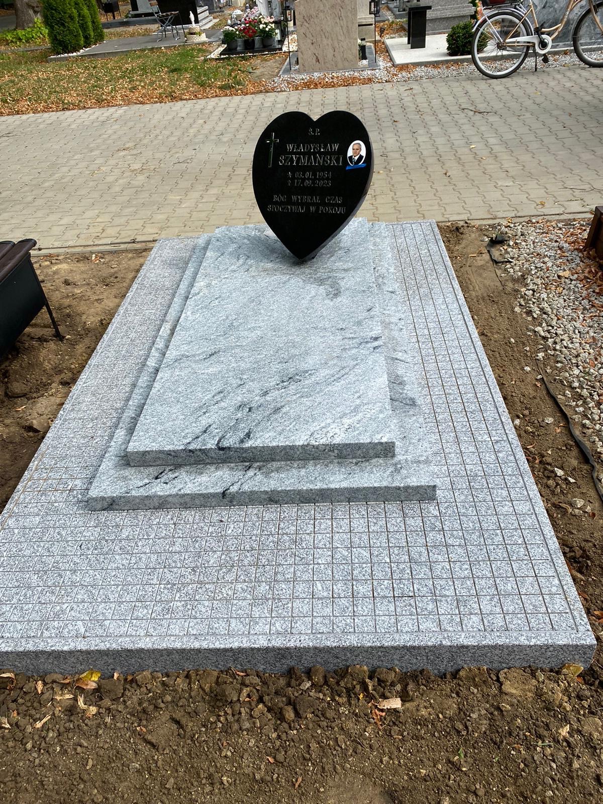 Granitowy nagrobek z czarnym sercem i portretem, otoczony kostką w kratkę. Widok na cmentarz, jesienne liście. Jasnoszary kamień, delikatne wzory.