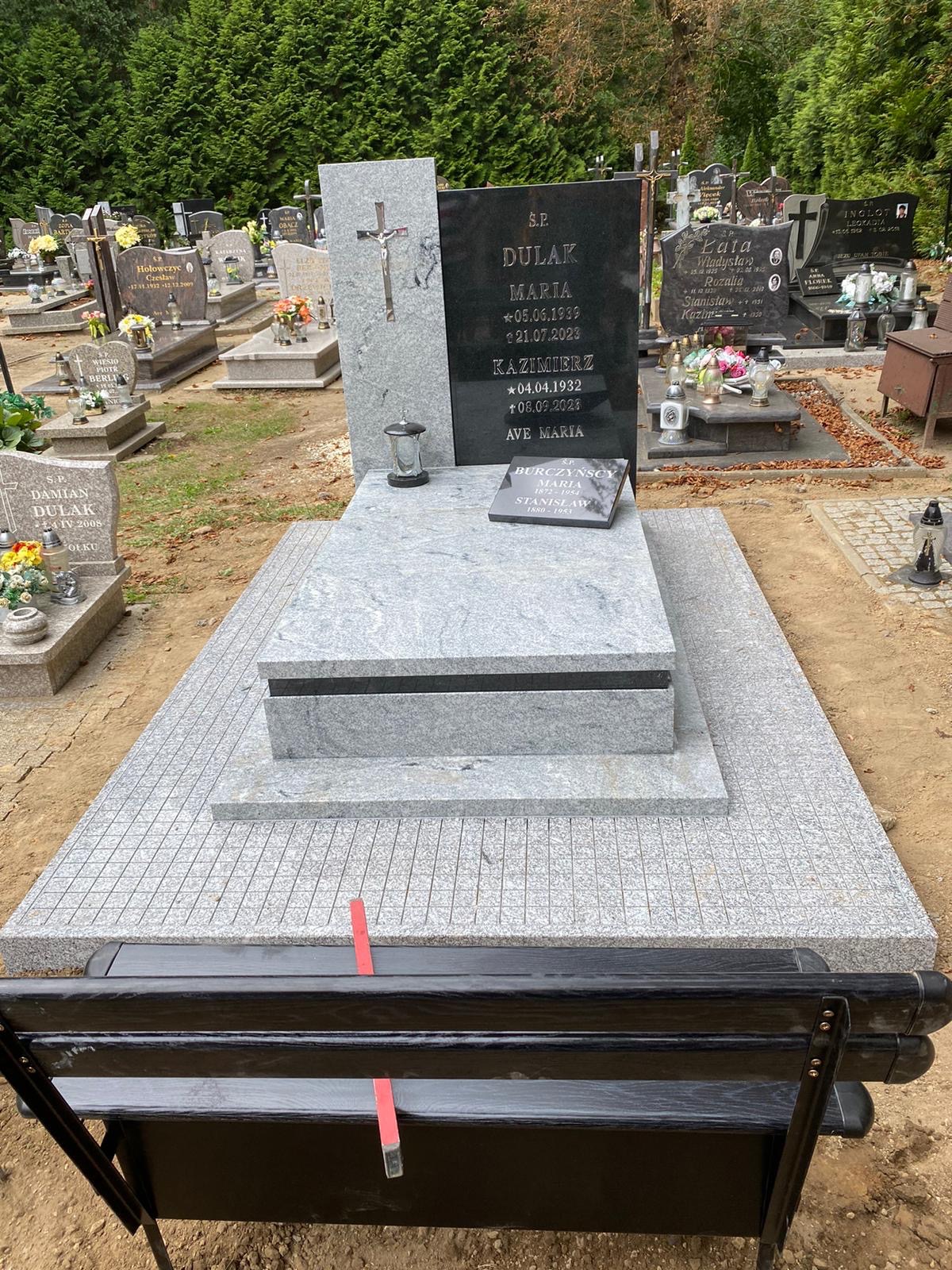 Granitowy nagrobek z czarną tablicą i krzyżem na cmentarzu. Jasnoszara podstawa z kostki. W tle inne groby i drzewa. Widoczna część czarnej ławki.