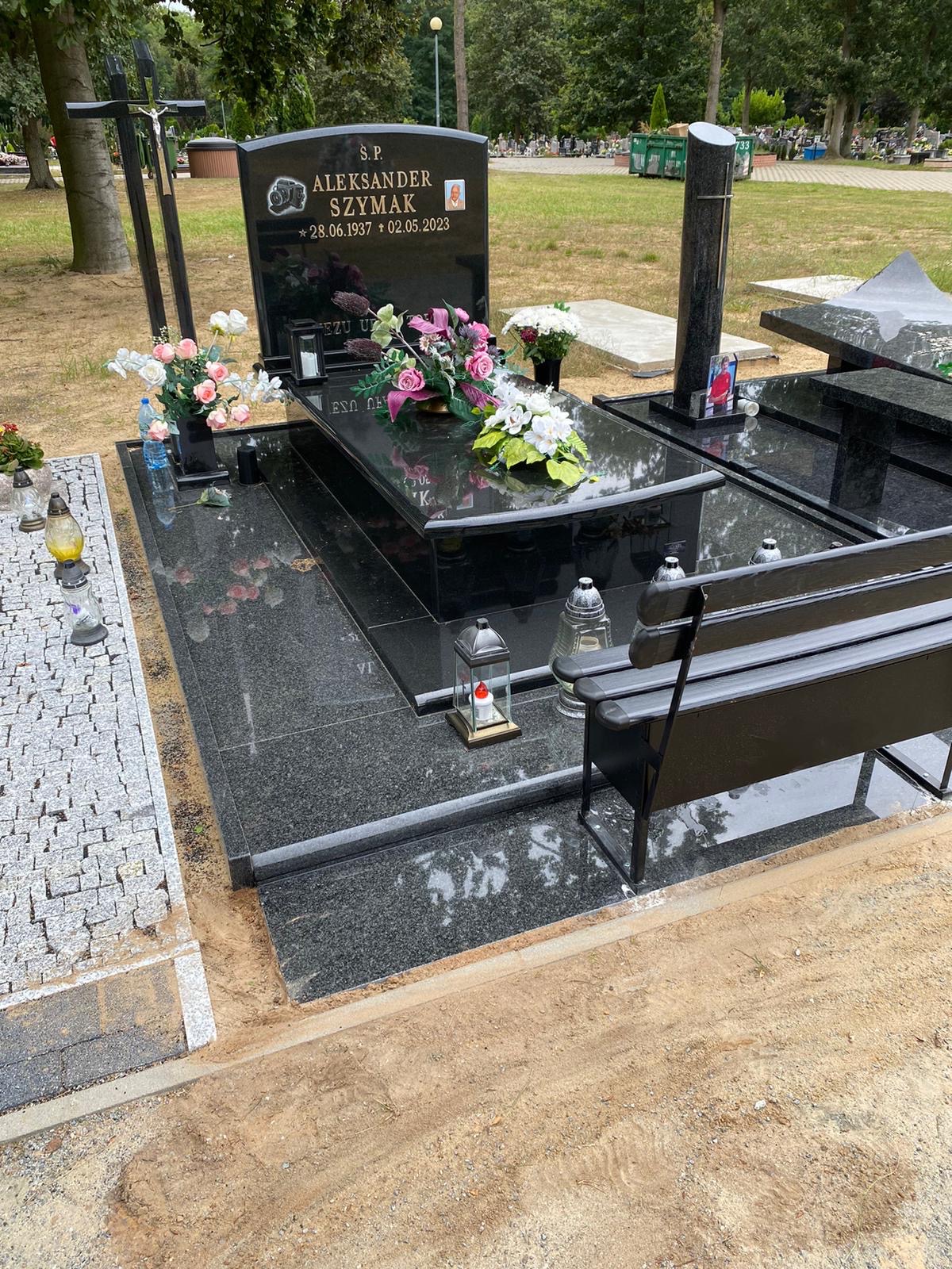 Czarny, wypolerowany nagrobek z granitu z kwiatami i ławką na cmentarzu. Widoczny krzyż, lampiony i woda mineralna. Otoczenie: drzewa, inne nagrobki.