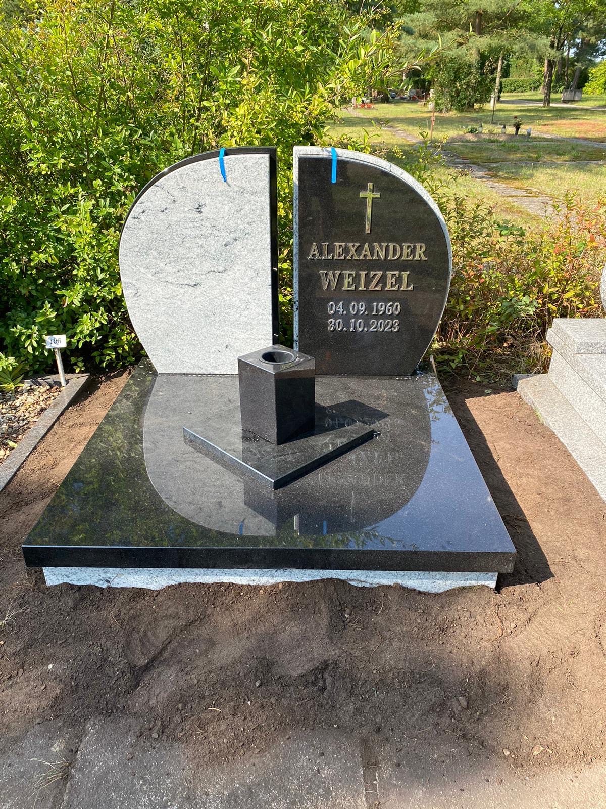 Nowoczesny, dwuczęściowy nagrobek z ciemnego granitu z wygrawerowanym imieniem i nazwiskiem oraz datami, otoczony zielenią na cmentarzu.