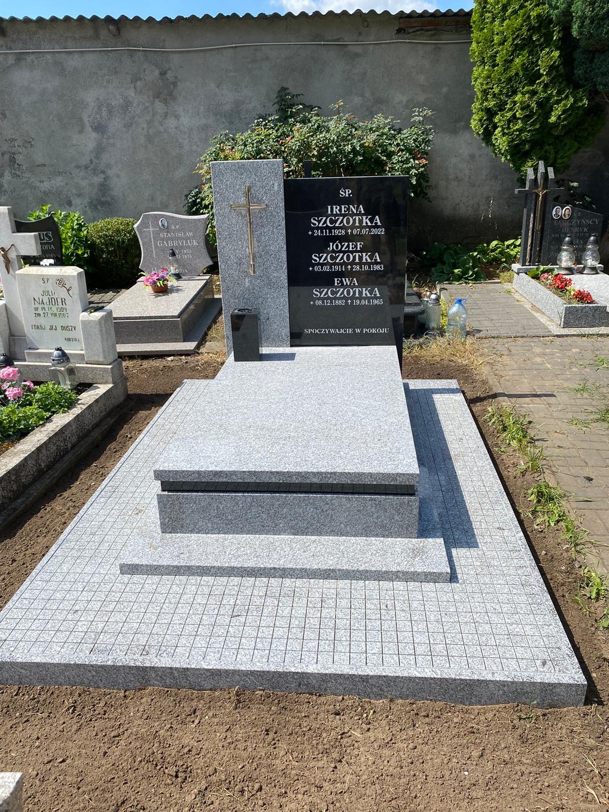 Granitowy nagrobek z czarną tablicą inskrypcyjną i złotym krzyżem, otoczony szarą kostką. Widok na cmentarz, zieleń i mur w tle.