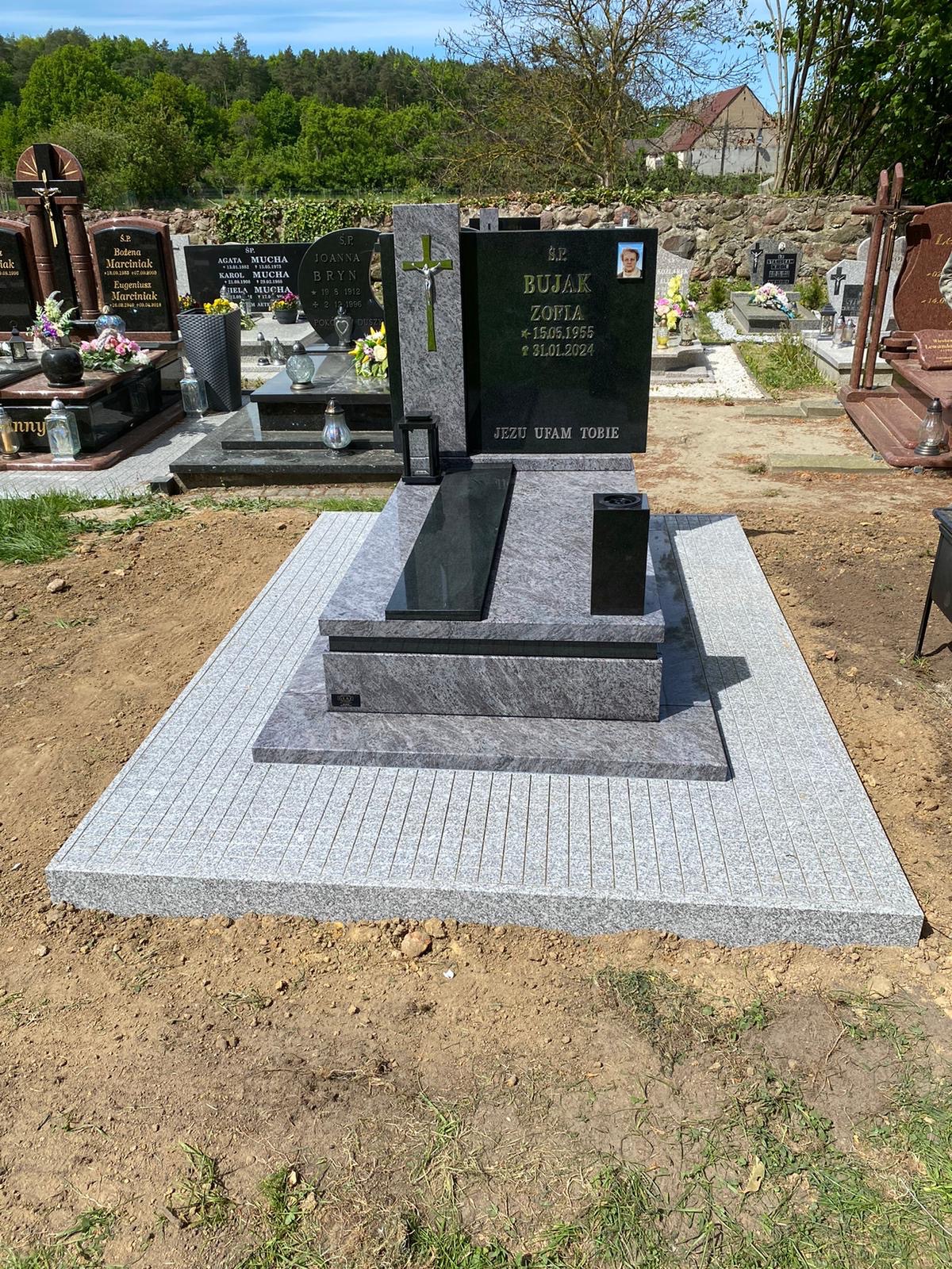 Granitowy nagrobek z ciemną tablicą i jasnym krzyżem, otoczony jasną kostką. Usługa kamieniarska w Pyrzycach. Widok na cmentarz i zieleń.