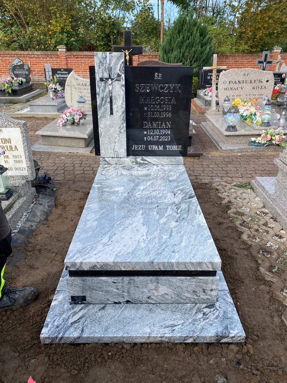 Granitowy nagrobek w odcieniach szarości z czarną tablicą i srebrnym krzyżem na tle cmentarza z innymi nagrobkami i zielenią.