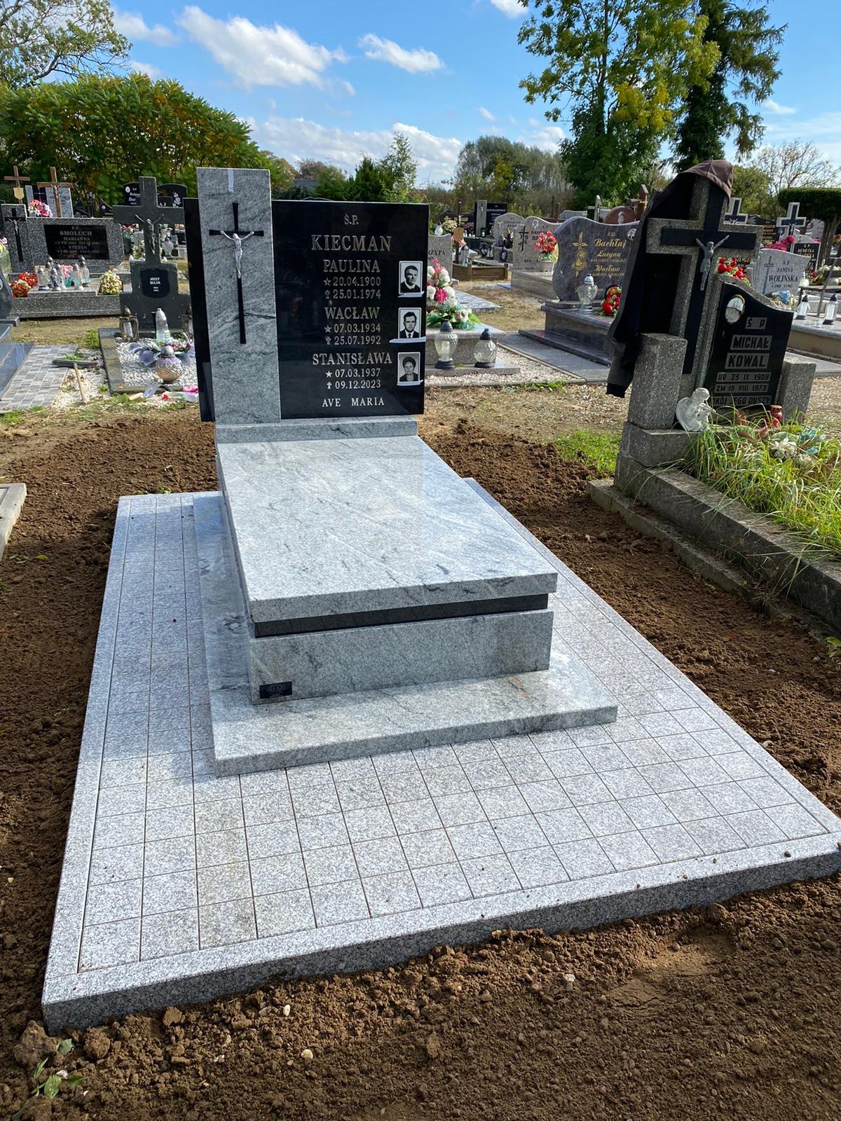 Jasnoszary, nowoczesny nagrobek z czarną tablicą i zdjęciami zmarłych. Otoczenie: cmentarz z innymi nagrobkami i zielenią. Całość w słoneczny dzień.