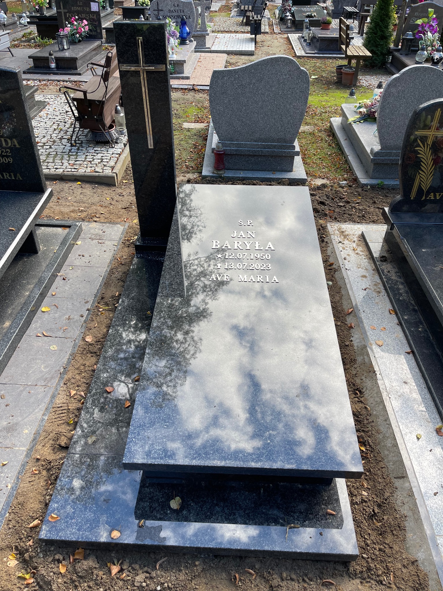 Granitowy nagrobek z inskrypcją 'S.P. Jan Baryła' i datami urodzenia oraz śmierci, otoczony ziemią. Na nagrobku odbija się niebo.
