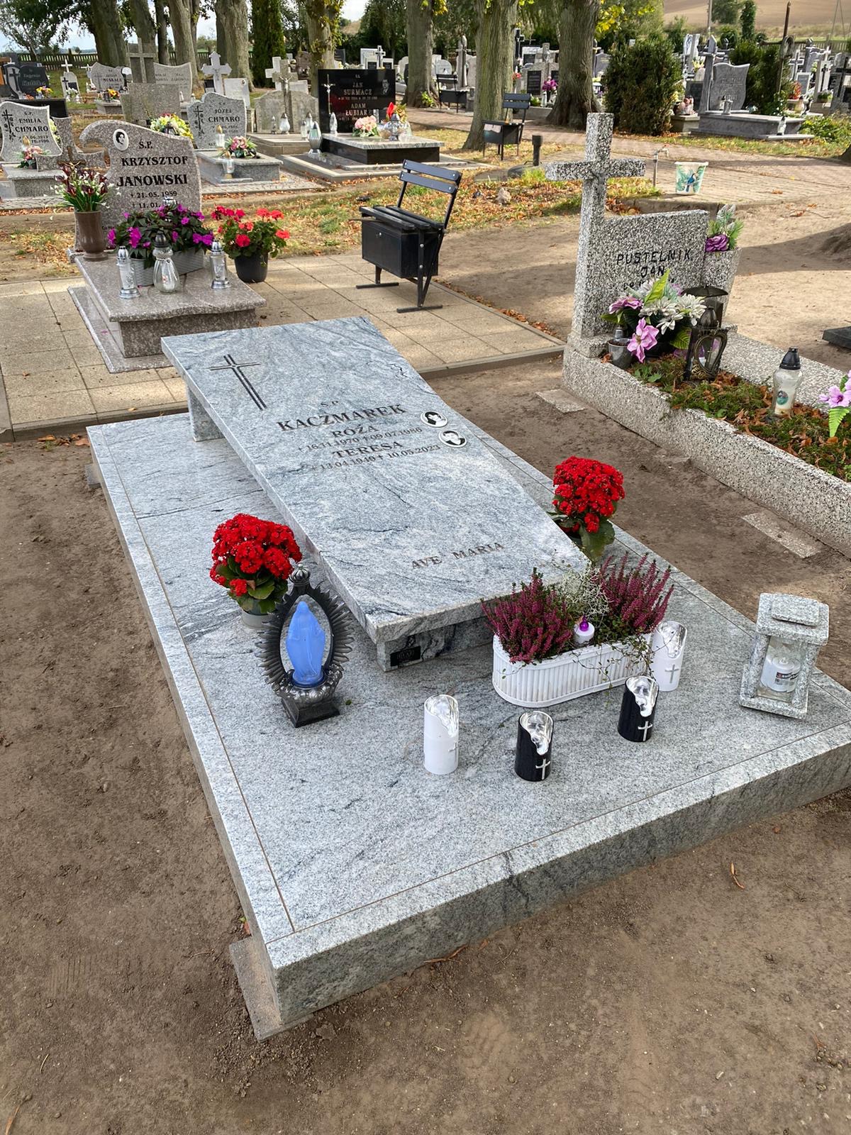 Granitowy nagrobek z inskrypcjami, kwiatami i lampionami na cmentarzu. Szara kolorystyka kamienia, krzyż na płycie. W tle inne nagrobki i drzewa.