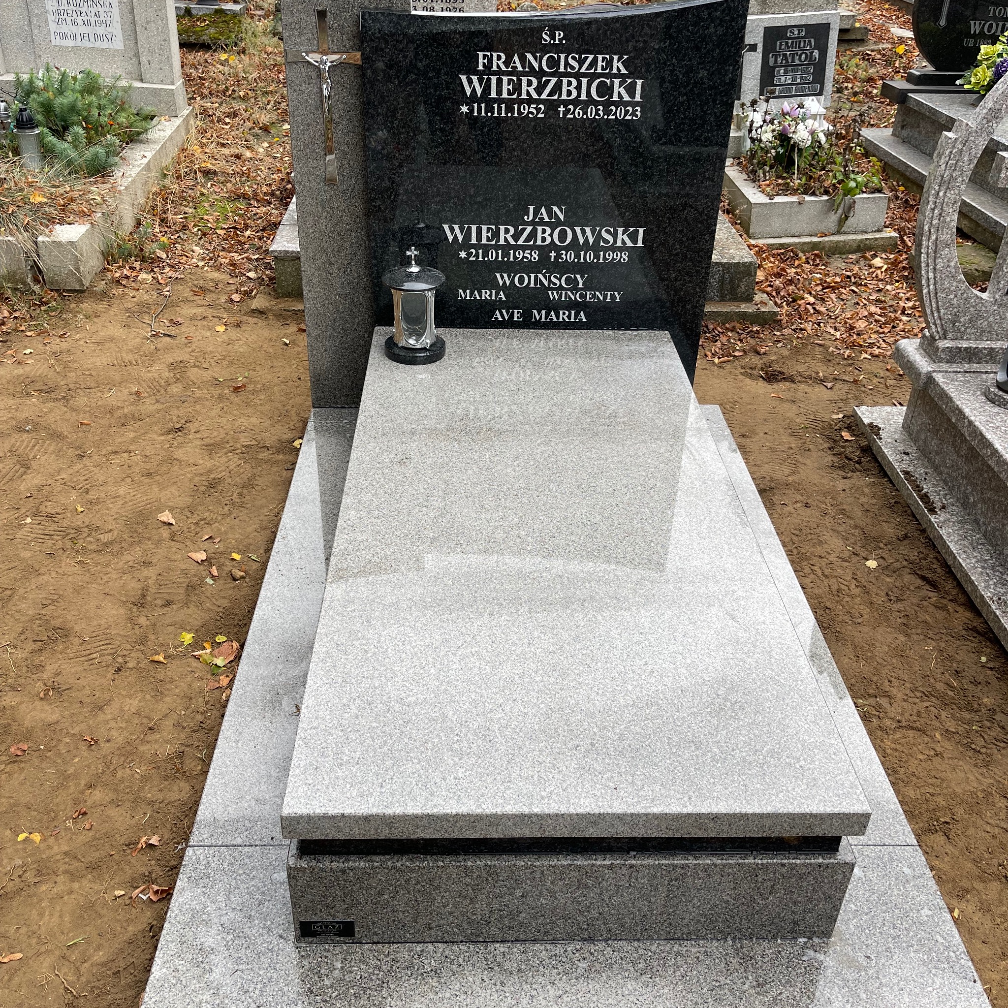 Granitowy nagrobek z czarną tablicą, krzyżem i lampionem. Jasnoszara płyta z napisem. Otoczenie cmentarza z liśćmi.