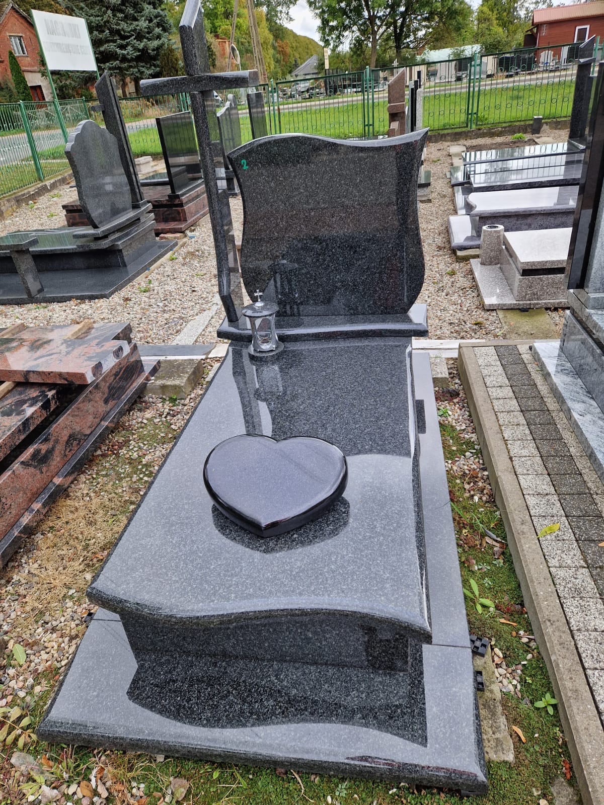 Czarny, granitowy nagrobek z krzyżem i sercem, ustawiony na zewnątrz, na tle innych nagrobków i zieleni. Widoczny lampion.