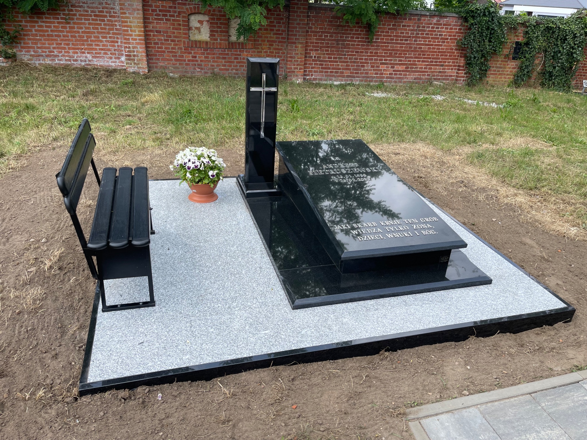 Czarny nagrobek z krzyżem i ławką na jasnoszarym podłożu, ozdobiony kwiatami w doniczce. W tle ceglany mur i zieleń.