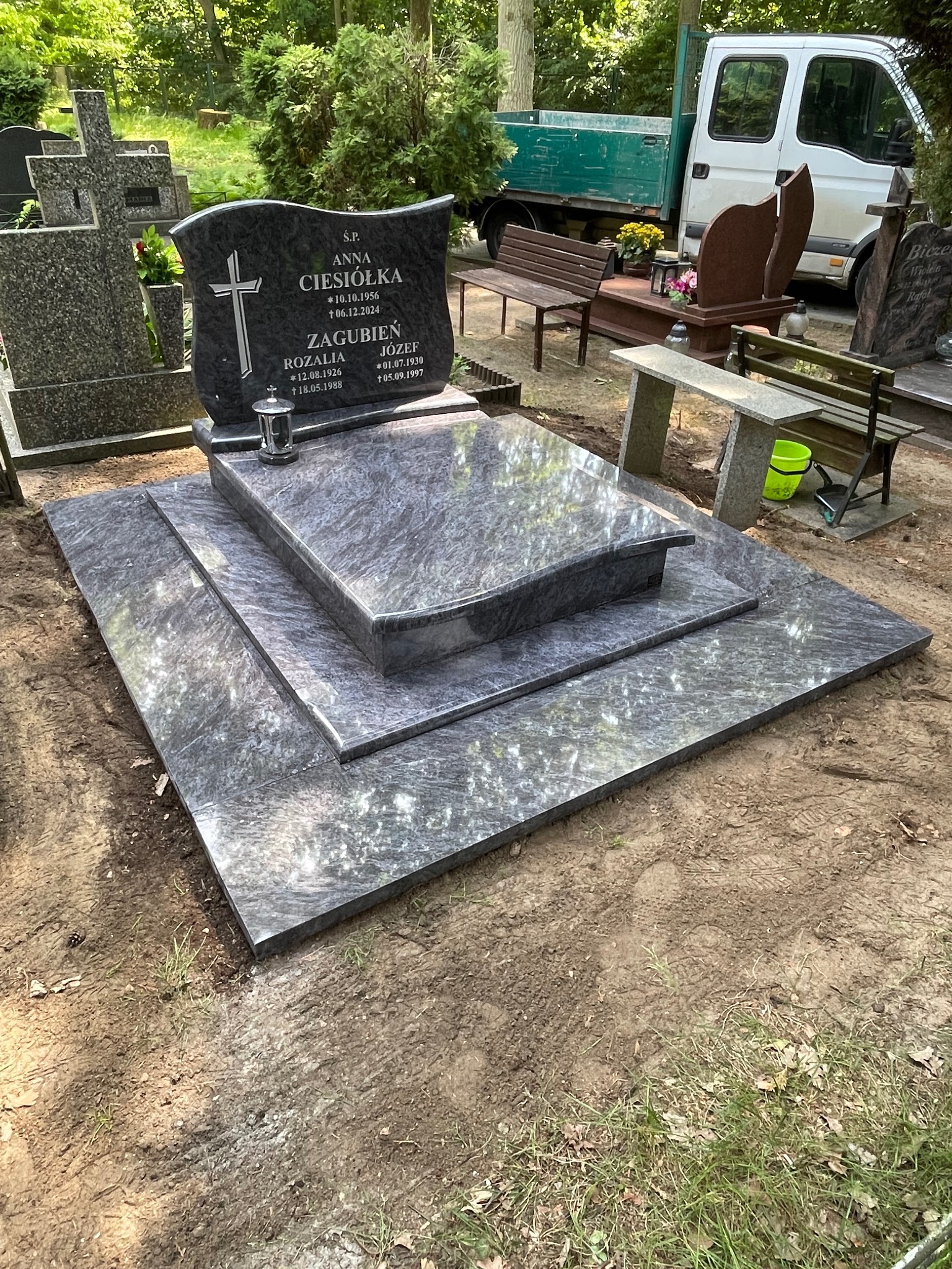 Granitowy nagrobek w odcieniach szarości z inskrypcją, otoczony ziemią i zielenią cmentarza. W tle widać biały samochód dostawczy i inne nagrobki.