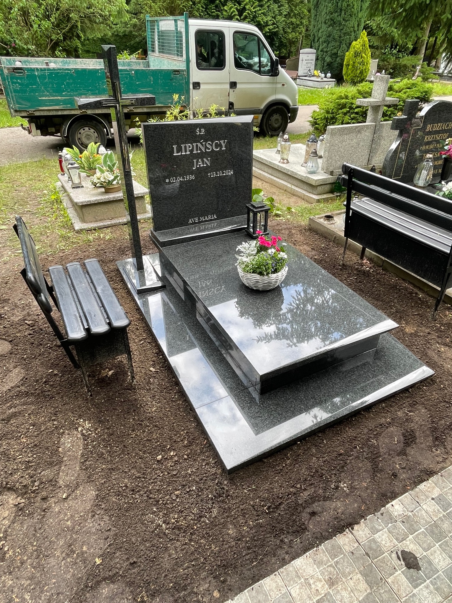 Granitowy nagrobek z napisem 'Lipinscy Jan' i datami, ozdobiony kwiatami w koszyku. W tle zielony samochód dostawczy i inne nagrobki na cmentarzu.