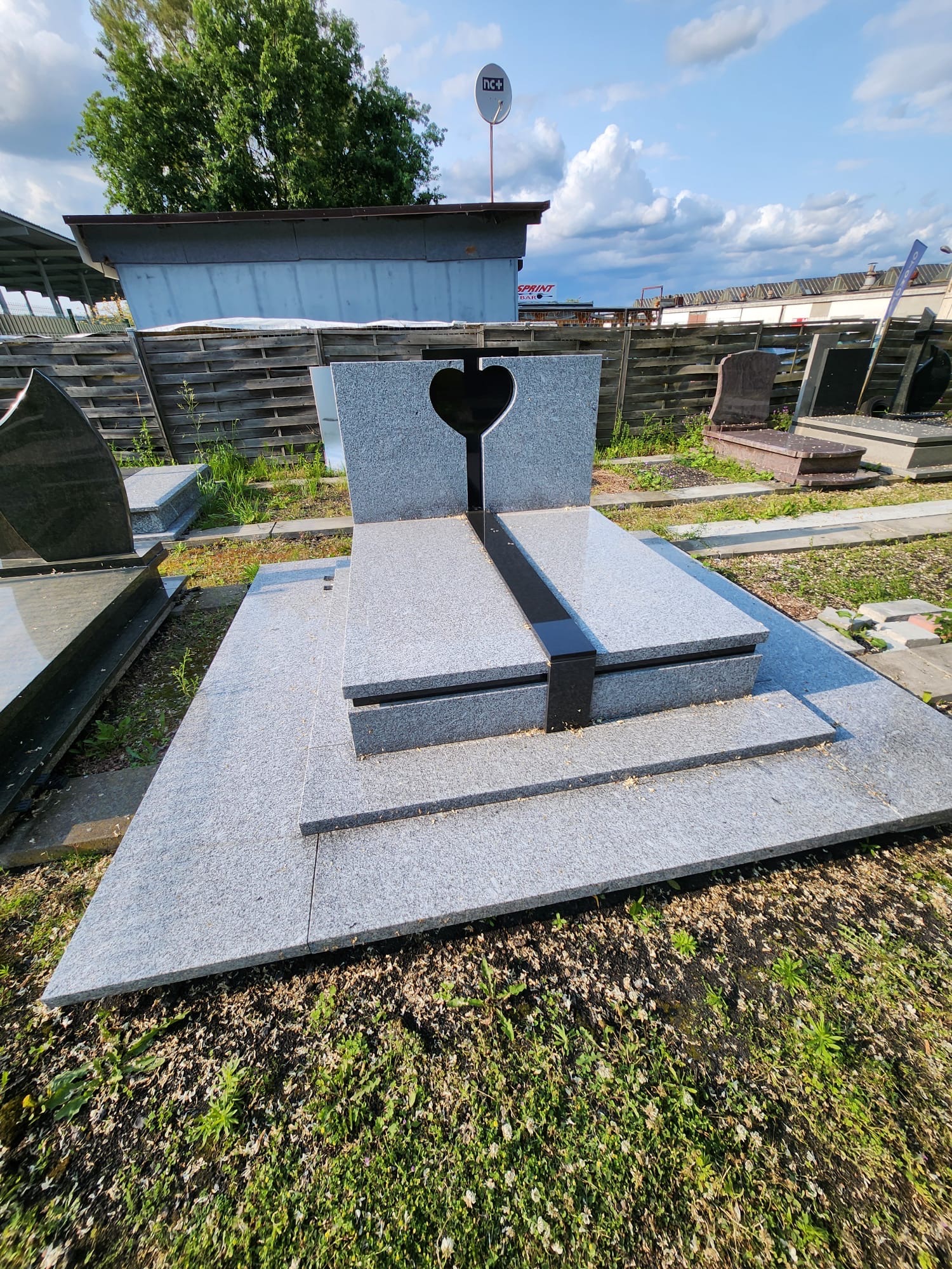 Jasnoszary nagrobek z wyciętym sercem, umieszczony na ekspozycji wśród innych nagrobków, w tle budynek i drzewa, widok z dołu.