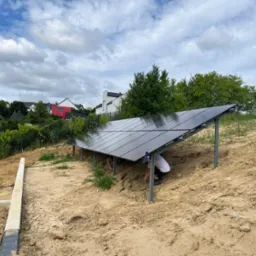 Montaż paneli słonecznych na konstrukcji wsporczej na nierównym terenie. Widoczni instalatorzy podczas pracy, błękitne niebo z chmurami w tle.
