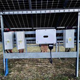 RicoSun Weronika Jankowska - Instalacja paneli słonecznych z widocznymi skrzynkami elektrycznymi i inwerterem na metalowej konstrukcji, na tle ziemi. Widoczne kable i oznaczenia ostrzegawcze.