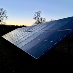 RicoSun Weronika Jankowska - Instalacja fotowoltaiczna na tle zachodzącego słońca. Panele odbijają niebo, widoczne drzewa bez liści i lampa uliczna. Ujęcie z dołu, podkreślające rozmiar instalacji.