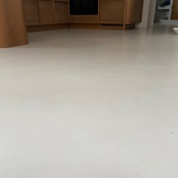 Micro Art Design - Jasna, gładka posadzka z mikrocementu w neutralnym odcieniu szarości, z widocznymi fragmentami zabudowy kuchennej i elementami wyposażenia wnętrza w tle.