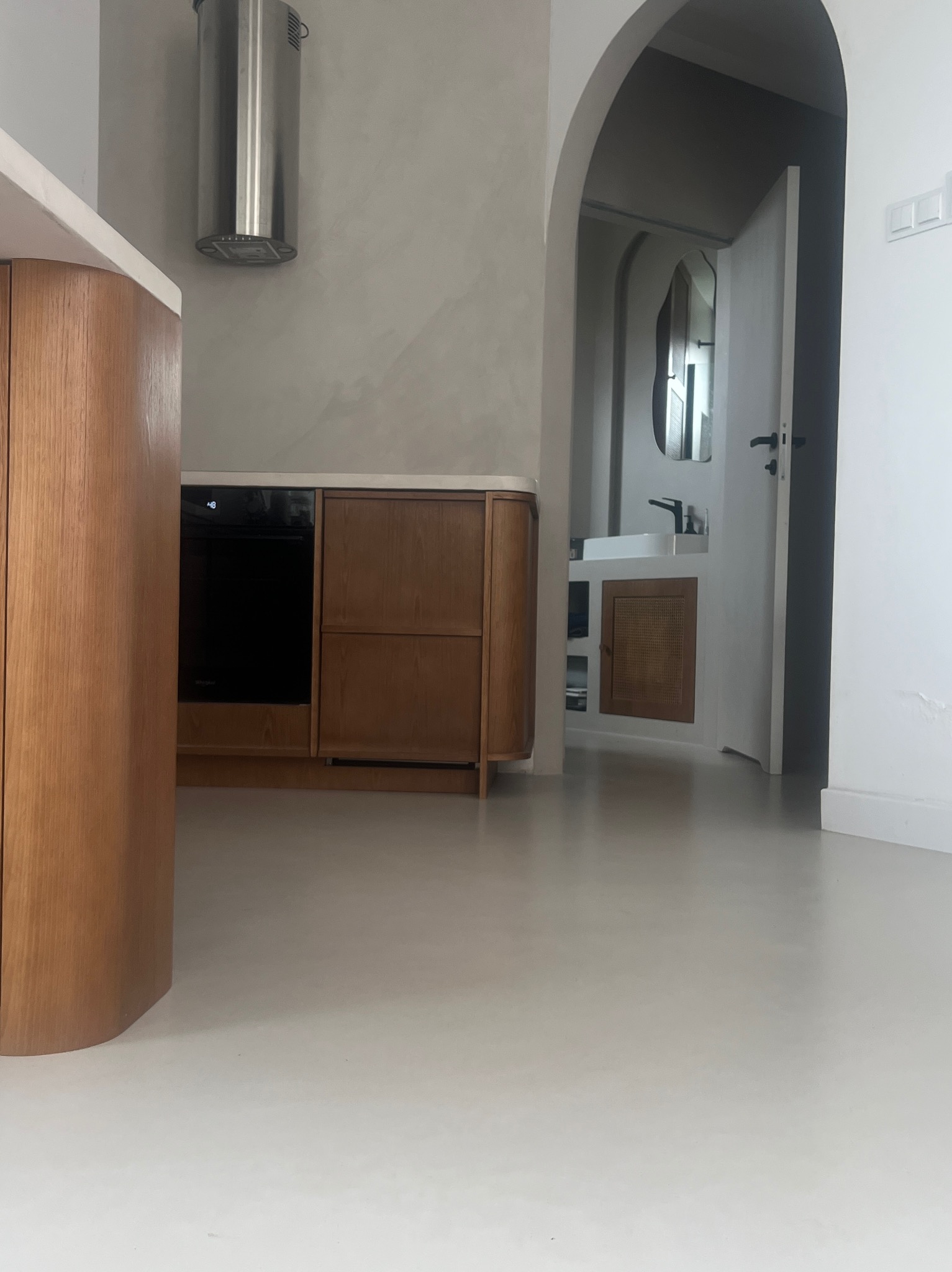 Jasna posadzka w minimalistycznej kuchni z drewnianymi meblami i łukowatym przejściem do łazienki. Gładka powierzchnia, neutralna kolorystyka.