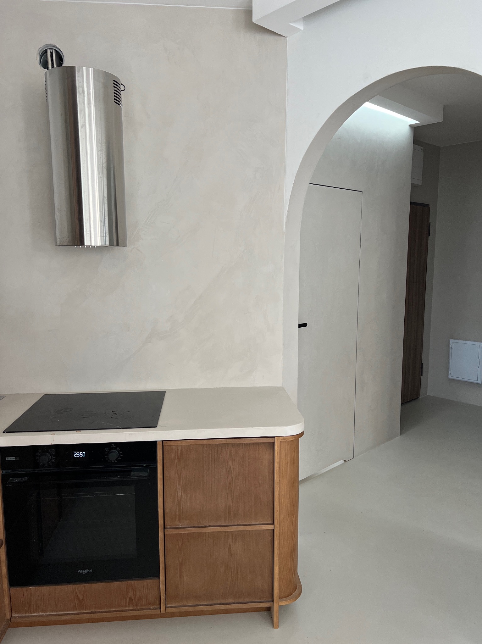 Kuchnia w neutralnych barwach z wykończeniem ścian przypominającym trawertyn, z łukowym przejściem do innego pomieszczenia i zabudową kuchenną z drewnianymi frontami.