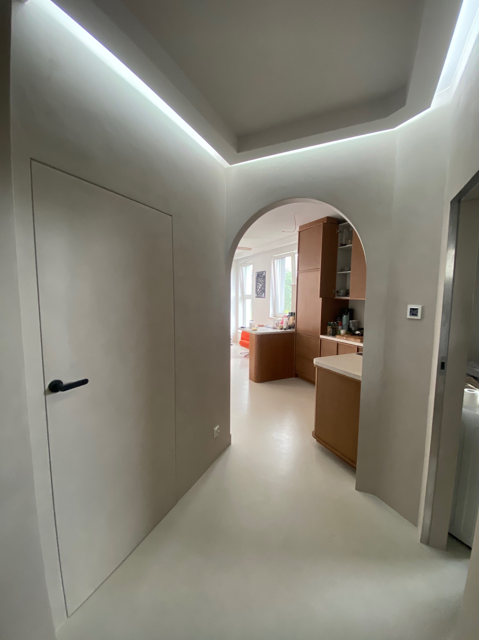 Minimalistyczne wnętrze z łukowym przejściem do kuchni. Białe ściany i podłoga, ukryte drzwi z czarną klamką, oświetlenie LED nad sufitem. Widok na drewnianą zabudowę kuchenną.