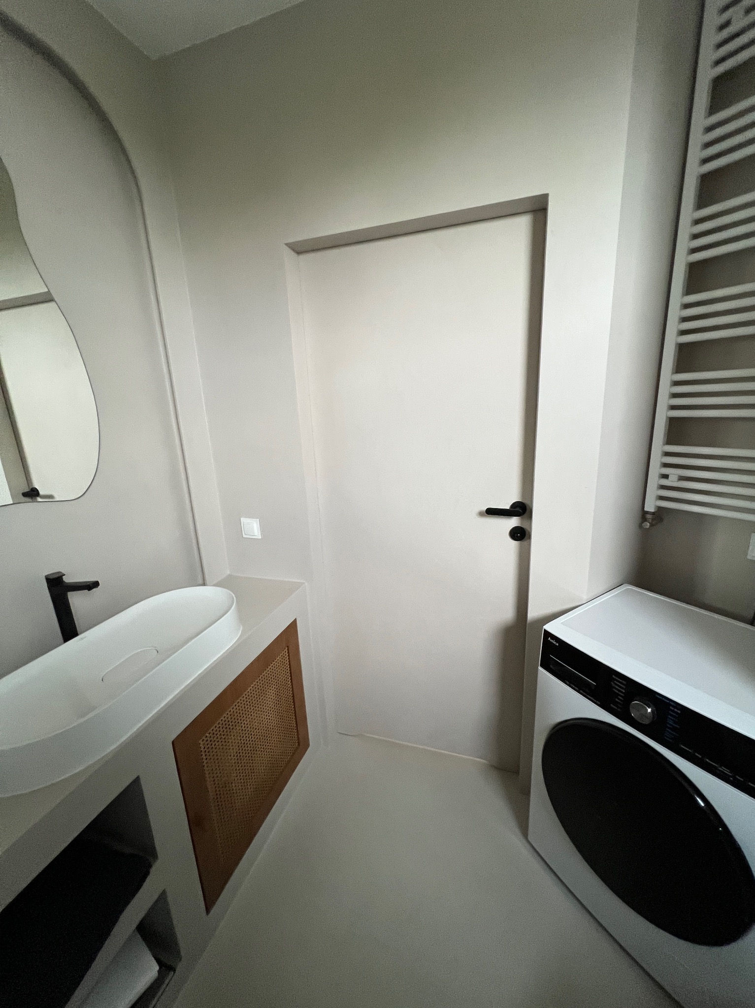 Beżowa łazienka z umywalką na blacie, drzwiami i pralką. Powierzchnie wykończone jednolitą, gładką powłoką. Minimalistyczny design.