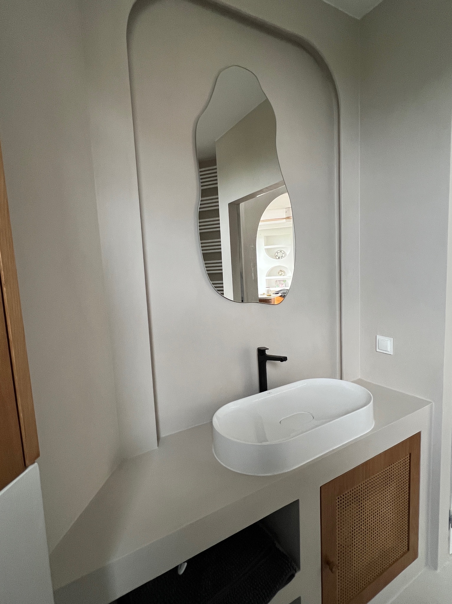 Beżowa łazienka z umywalką nablatową i asymetrycznym lustrem w pasującej wnęce. Widoczna szafka z rattanowymi drzwiczkami i czarna bateria.