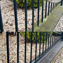 MOBIL WASH BARTEK WALCZEWSKI - Fragment czarnego, metalowego ogrodzenia po czyszczeniu, z widoczną różnicą w czystości na betonowym murku. Kamienie dekoracyjne w tle.