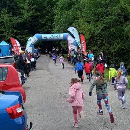PYCHOTKA CAFE Jakub Pych - Bieg dziecięcy na asfalcie, z bramą startową i kibicami. W tle las, pochmurne niebo. Widok z perspektywy uczestnika, dynamika ruchu.