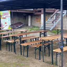 PYCHOTKA CAFE Jakub Pych - Ustawione rzędy drewnianych stołów i ławek pod czarnym namiotem, gotowe na imprezę plenerową. W tle betonowy mur z reklamą i schody.