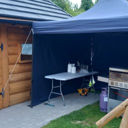 PYCHOTKA CAFE Jakub Pych - Rozłożony namiot cateringowy z grillem, stolikiem i butlą gazową obok drewnianego domku, przygotowany do obsługi imprezy plenerowej. Widok z boku, na trawie.