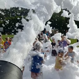 PYCHOTKA CAFE Jakub Pych - Dzieci bawią się w pianie podczas imprezy plenerowej. Duży strumień piany zalewa grupę roześmianych dzieci. Widoczne drzewa i niebo w tle. Zabawa z animatorem.