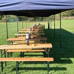 PYCHOTKA CAFE Jakub Pych - Ustawione stoły i ławy pod namiotami na zielonej trawie, gotowe na imprezę plenerową. Na stołach woda i przekąski. W tle drzewa i budynki.