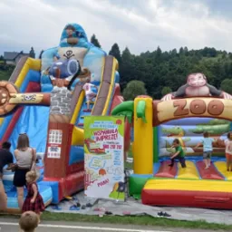 Dmuchane zamki: piracki zamek ze zjeżdżalnią i zamek zoo z małpą. Dzieci bawią się na dmuchańcach podczas imprezy plenerowej. W tle zielone wzgórza.