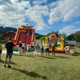 Plenerowa impreza z dmuchańcami na trawie, w tle góry i niebo z chmurami. Widoczna scena, grupa ludzi, zjeżdżalnia i dmuchany zamek.