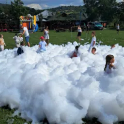 Dzieci bawią się w pianie na zielonym trawniku, w tle dmuchany zamek i budynki. Aktywna zabawa na świeżym powietrzu podczas imprezy plenerowej.