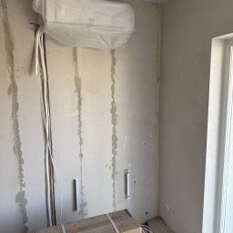 ISCOLD Dennis Bauwens - Montaż klimatyzacji Sinclair w surowym wnętrzu. Jednostka wewnętrzna w folii ochronnej, kable i rury prowadzone po ścianie, karton na podłodze. Prace instalacyjne w toku.