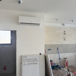 ISCOLD Dennis Bauwens - Biała klimatyzacja Sinclair na ścianie w surowym wnętrzu. Widoczne materiały budowlane, płytki i narzędzia. W tle ściana w trakcie wykończenia.