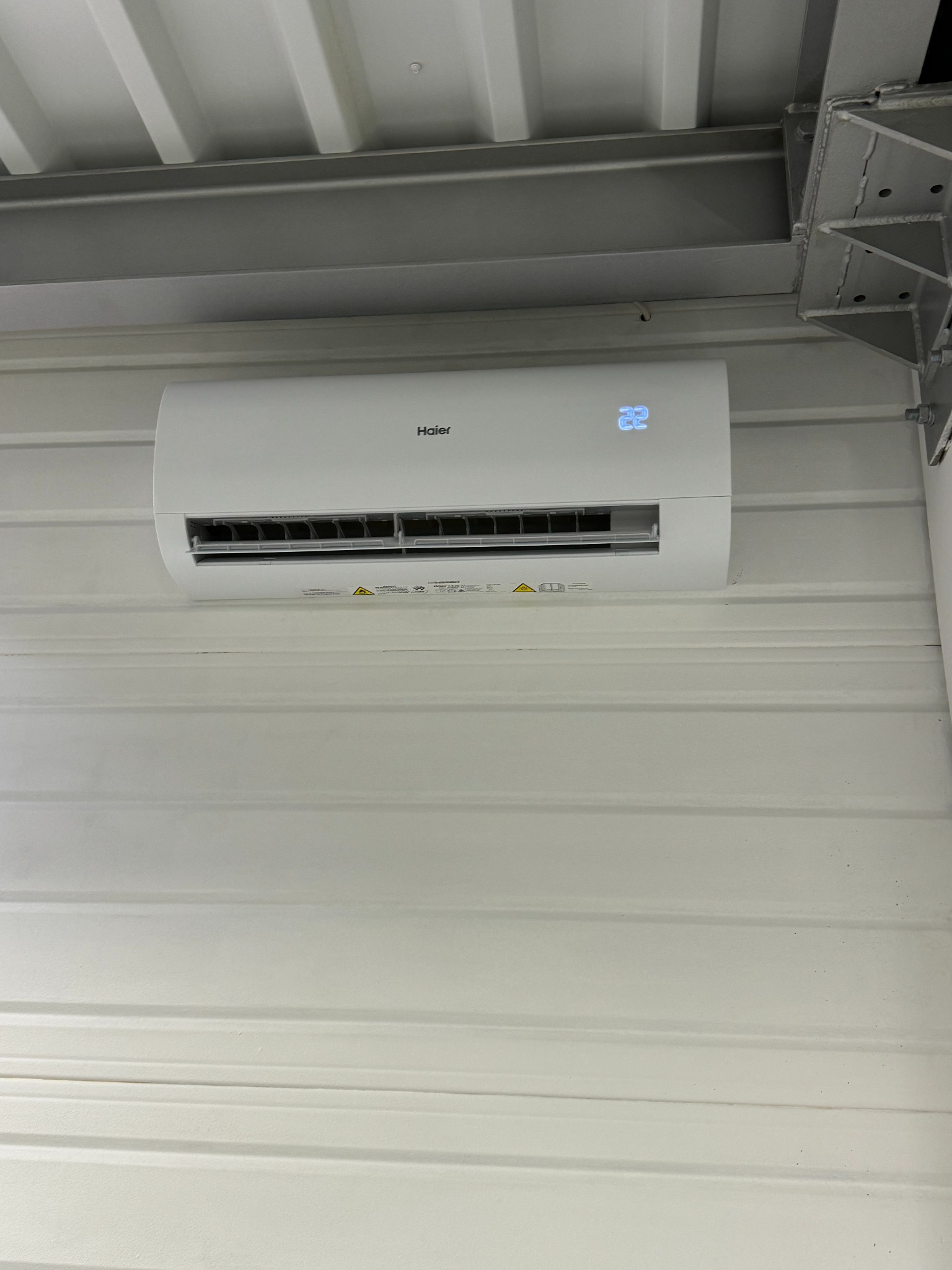 Biały klimatyzator Haier zamontowany na ścianie z blachy trapezowej, widoczny wyświetlacz z temperaturą 22 stopnie, oświetlenie górne.