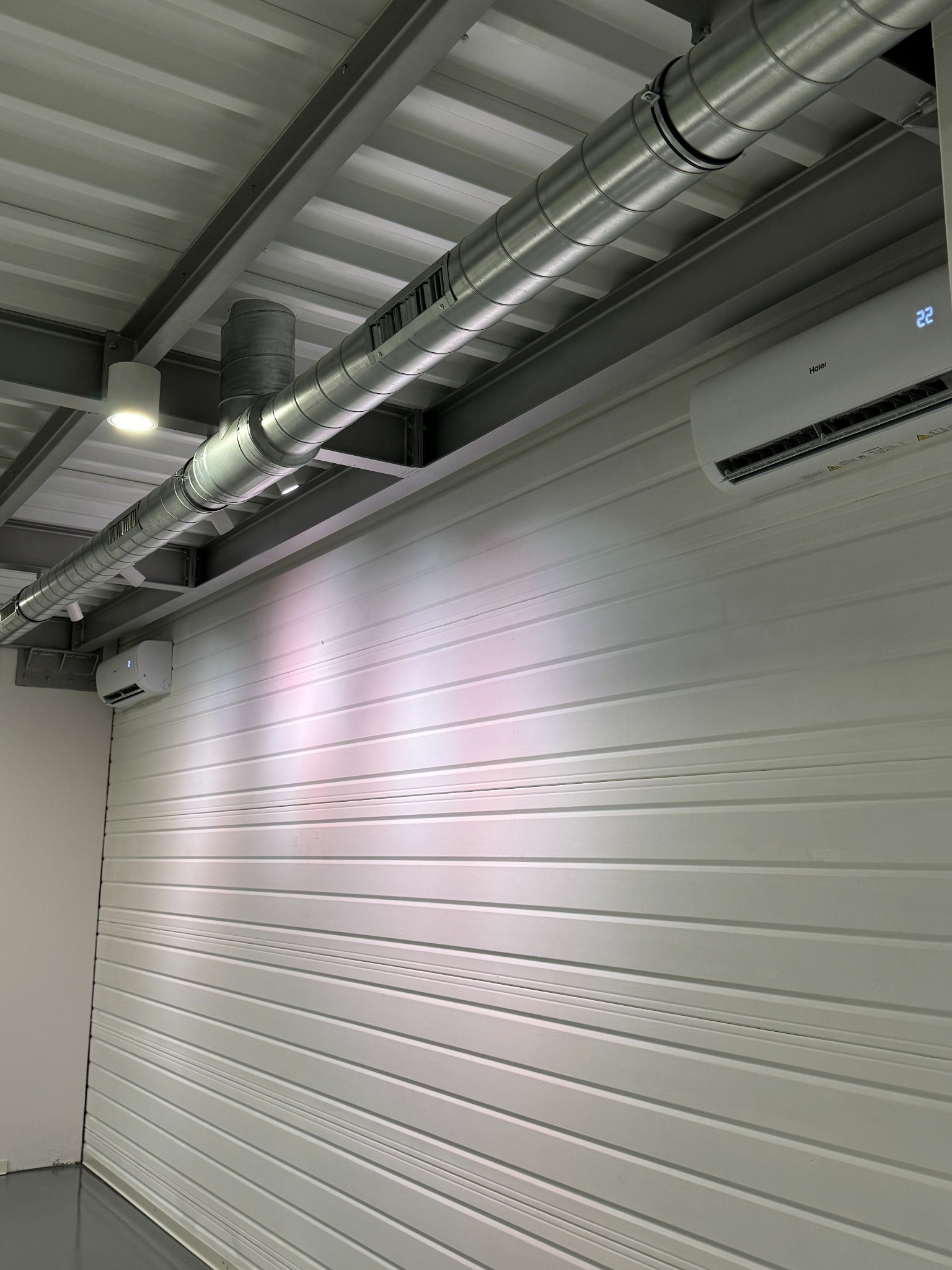 Wnętrze hali z zamontowaną klimatyzacją Haier i systemem wentylacyjnym z rurami pod sufitem. Widoczny fragment ściany z blachy trapezowej i oświetlenie.