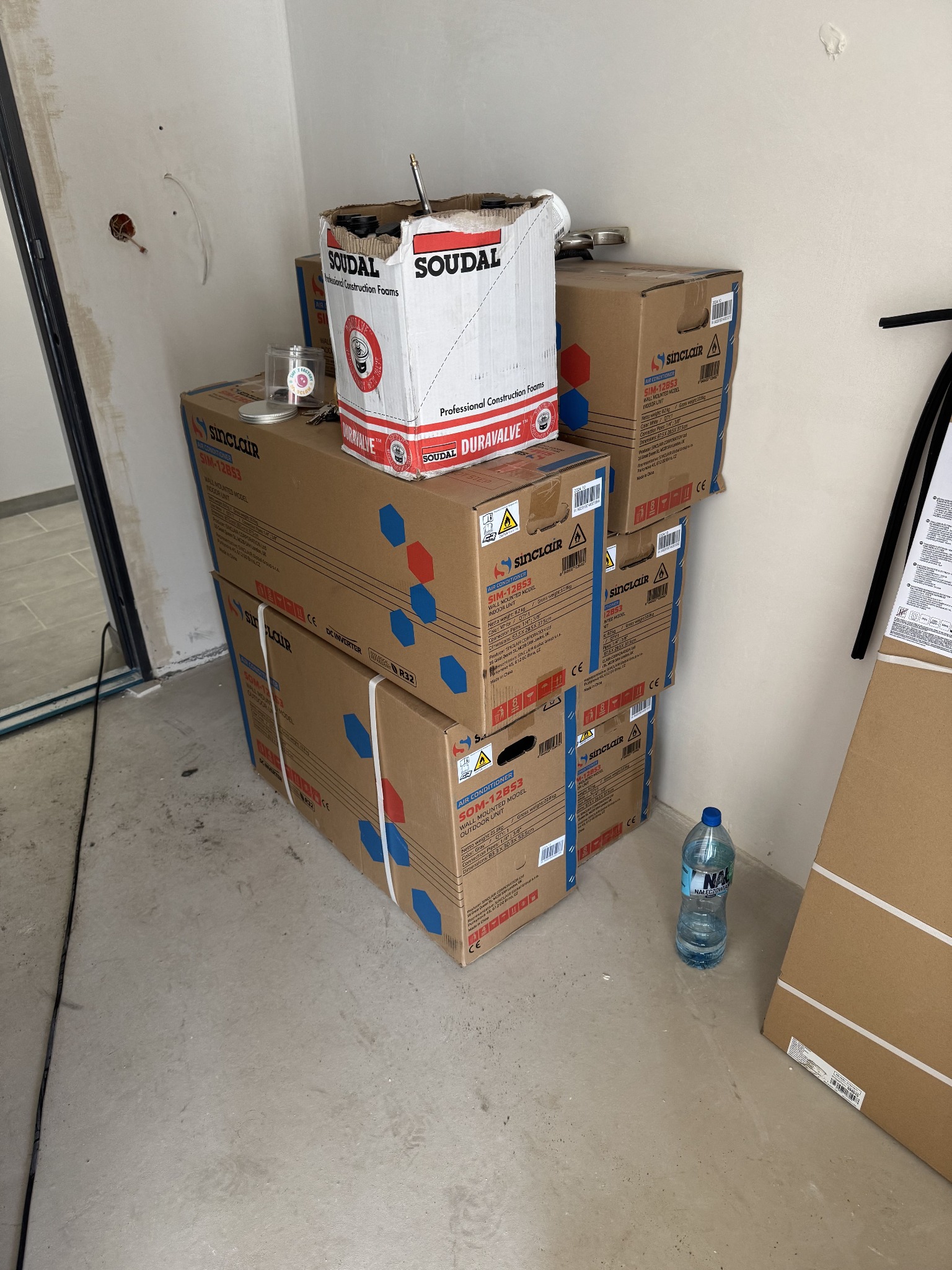 Kartonowe opakowania klimatyzatorów Sinclair SIM-12B53 i puszka pianki montażowej Soudal Duravalve na podłodze w surowym wnętrzu, butelka wody obok.