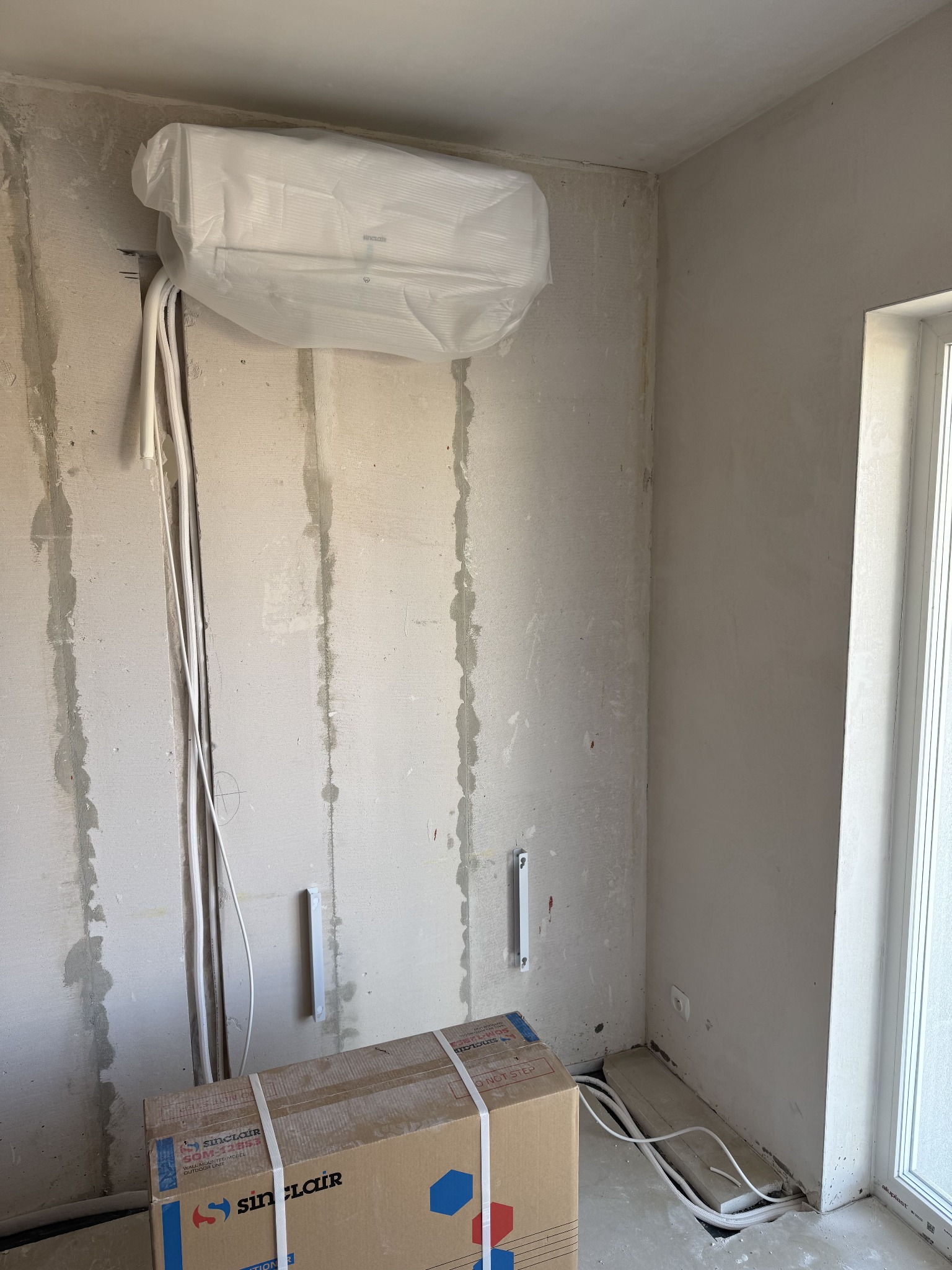 Montaż klimatyzacji Sinclair w surowym wnętrzu. Jednostka wewnętrzna w folii ochronnej, kable i rury prowadzone po ścianie, karton na podłodze. Prace instalacyjne w toku.