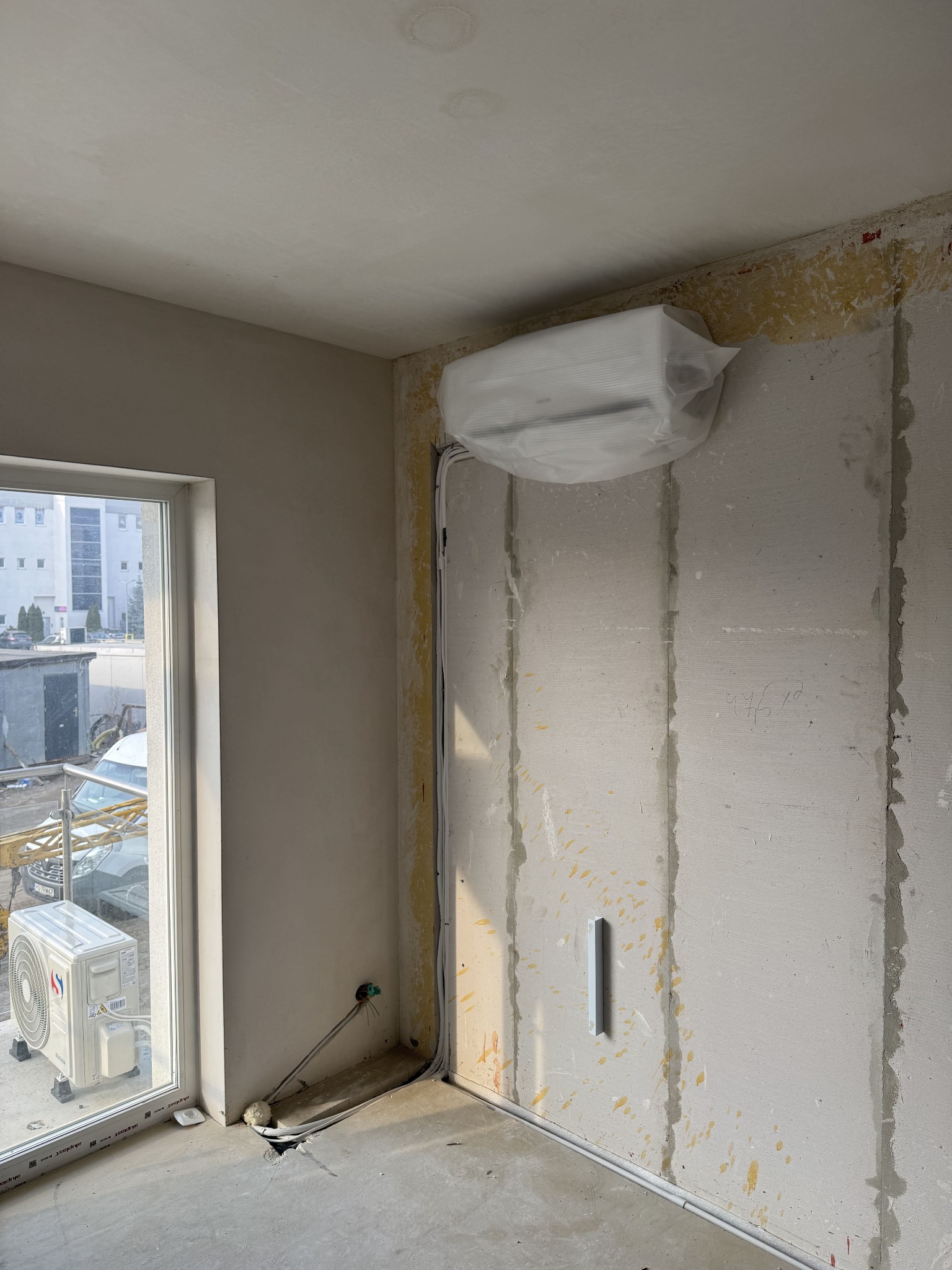 Wnętrze w trakcie montażu klimatyzacji. Jednostka wewnętrzna zabezpieczona folią, jednostka zewnętrzna widoczna za oknem. Ściany w surowym stanie, widoczne przewody.