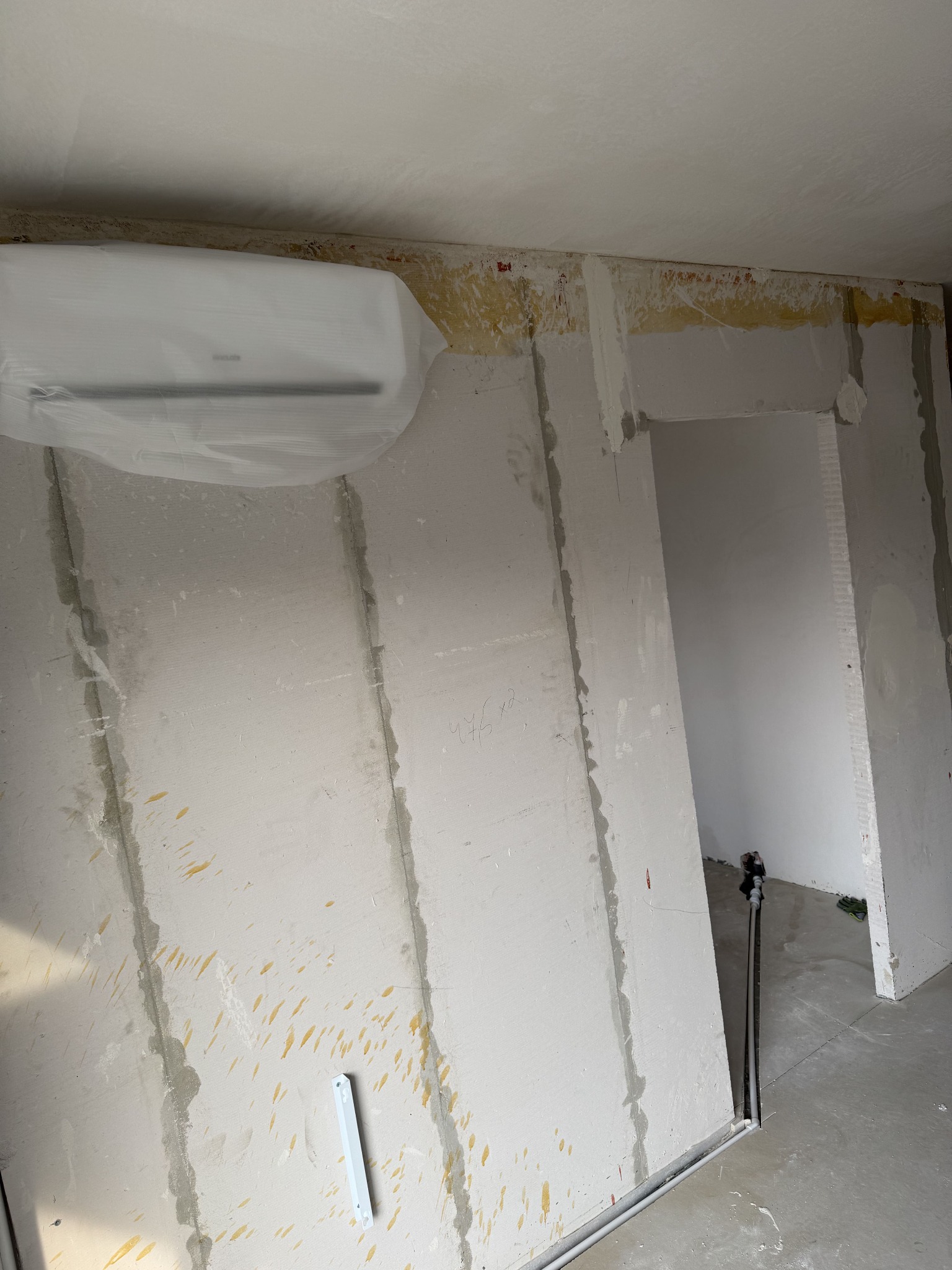 Klimatyzator w folii ochronnej, zamontowany na surowej ścianie w trakcie remontu. Widoczne ślady po pracach budowlanych i instalacje.